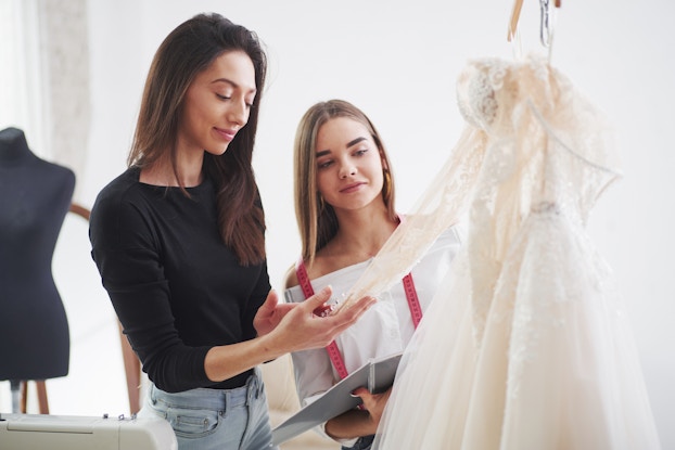  Two women examine a wedding dress hanging on a wooden hanger. The dress is a white off-the-shoulder gown with long semi-transparent sleeves, a lace-embroidered bodice, and a long tulle skirt. The woman on the left is taller with long dark hair; she holds the end of one of the sleeves between her hand, as if to feel the fabric. The woman on the right is shorter with long dark blonde hair; she has a red measuring tape around her neck and holds an open notebook.