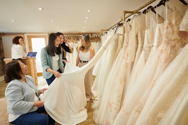 A bride-to-be is smiling as she looks at a wedding dress hanging on a rack in a bridal store. She is accompanied by her mother and friends. She is excited because she found "the dress."