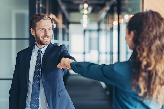 A man in businesswear, facing the camera, fist bumps a woman facing away from the camera. The background is an out-of-focus hallway with glass doors.