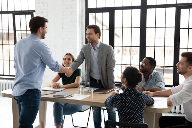  A group of employees in business casual clothes sit and stand around a wooden table in a large room. Two of the men at the table are standing and shaking hands; the rest of their coworkers look up at them and smile.