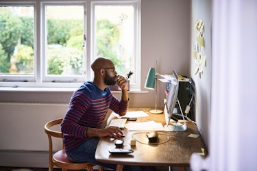  A wide shot of a bearded man sitting at a desk, looking at a computer monitor. The man is wearing glasses and a blue shirt with orange horizontal stripes. He has one hand to his mouth in a thoughtful expression and the other hand on a computer mouse. The desk also holds a lamp, a cordless keyboard, a cordless phone, a smartphone, and a stacked set of trays holding various papers. 