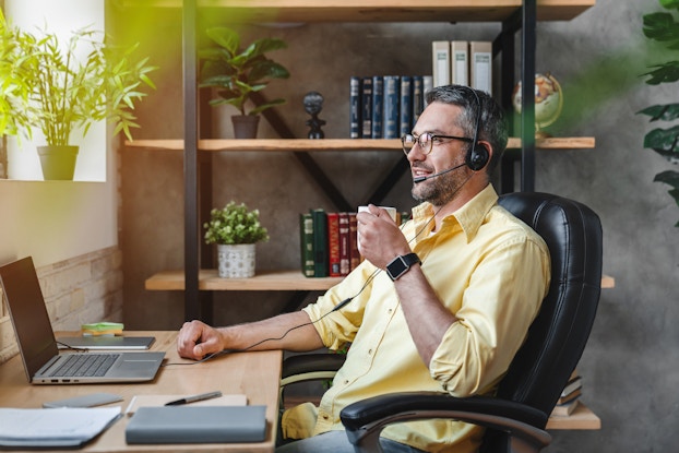  A man with graying hair and a stubbled chin sits at a desk, holding a mug and wearing a wired headset. He has a cup of coffee in his hand and gazes at the window above the desk with a slight smile. The wire on his headset connects to a laptop on the desk. The desk also holds a book, a stack of Post-It notes, a couple of stacks of paper, and a pen, all neatly arranged.