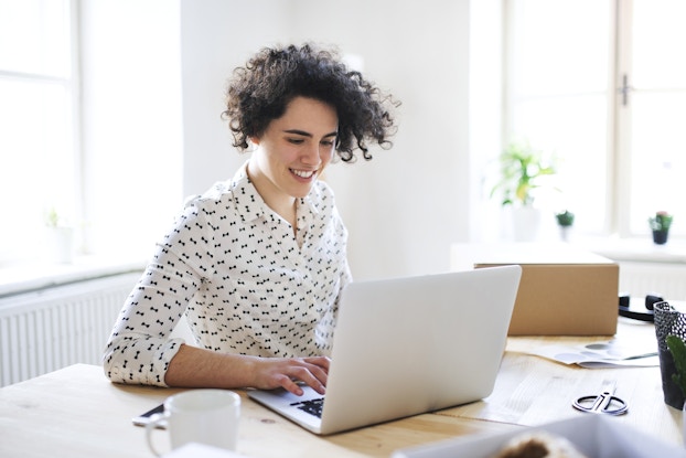 A curly-haired woman sits at a table and types on a laptop. The woman is smiling and wearing a white button-up shirt covered with small bow-shaped designs. The room in the background is white-walled with large windows letting in light from outside.