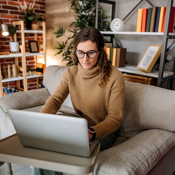 A woman wearing glasses and a tan turtleneck sweater sits in a plush beige armchair and looks at an open laptop. The laptop sits on a small plastic table in front of the chair. In the background are several shelves filled with books, framed pictures, and a few potted plants.