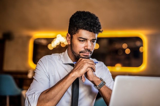 A mid-adult man is focused on reading a white paper on his laptop computer.
