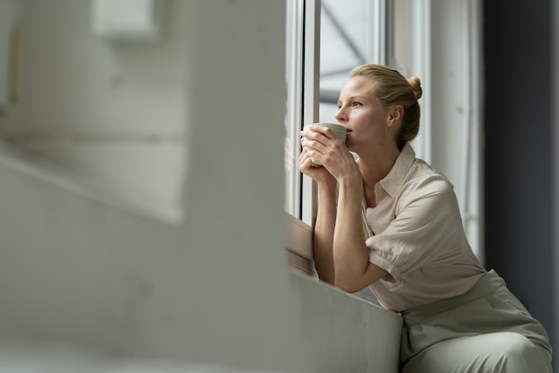  Woman in business clothes looking out the window while holding a coffee cup.