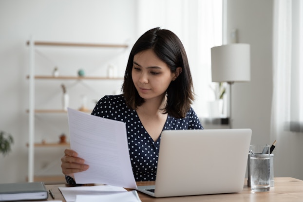 A young woman considers the contents of a piece of paper as she sits in front of a laptop.