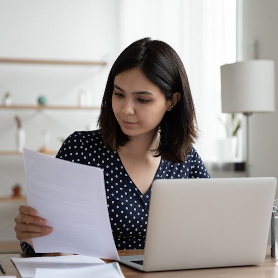 A young woman considers the contents of a piece of paper as she sits in front of a laptop.