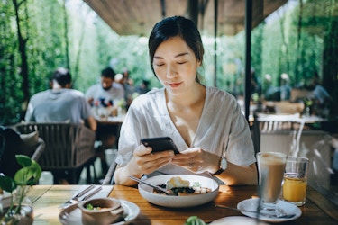  Smiling young woman using smartphone while having brunch in an outdoor restaurant surrounded by foliage and beautiful sunlight. 