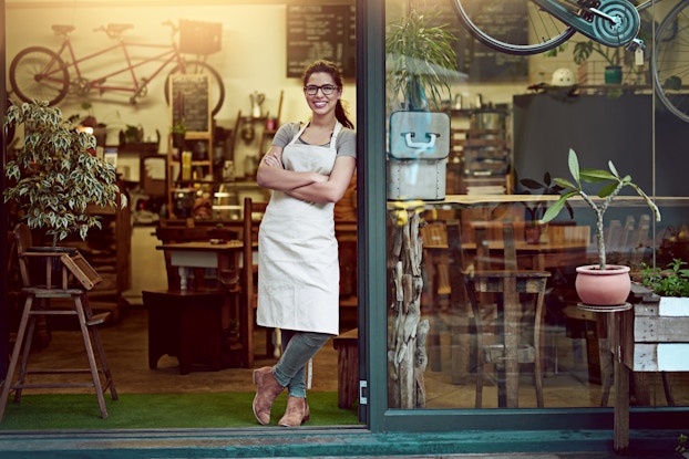 A woman in an apron stands in the doorway of a shop. Through the door and a floor-to-ceiling window can be seen wall-mounted bikes, wooden tables and chairs, decorative plants and a blackboard menu.