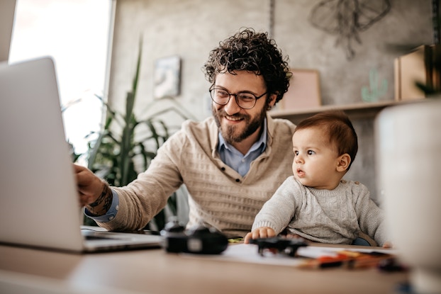  Man working from home on laptop while holding a baby on his lap.