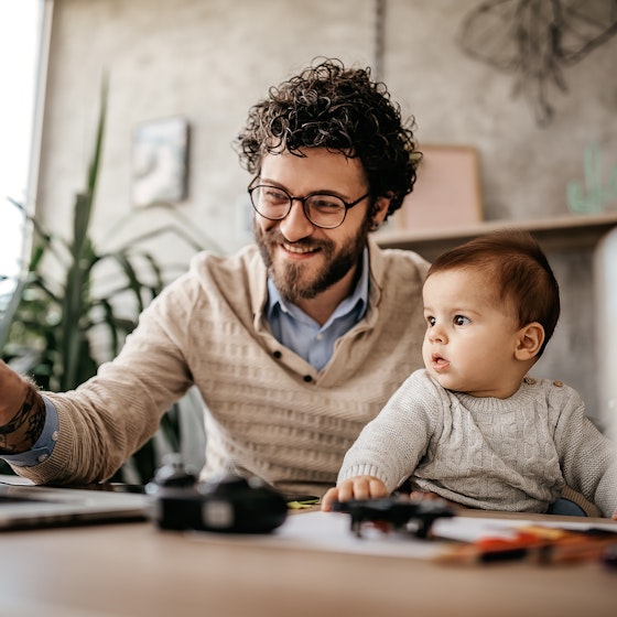 Man working from home on laptop while holding a baby on his lap.
