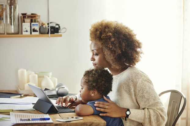 A woman sits at a table scattered with papers and works on a smart tablet with an attached keyboard. Her child, a toddler, is seated in her lap.