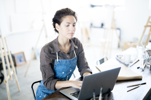  A woman in an artist's studio at a table, typing on a laptop. The woman has short, dark hair, and she wears a stained denim apron over a dark gray button-up shirt. In the background are several wooden easels and blank canvases.