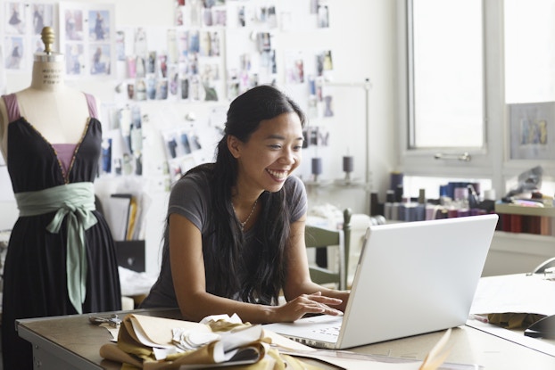 A smiling woman leans on a table in a fashion designer's studio and types on an open laptop. Beside the laptop are folded lengths of fabric in various shades of brown and white. Behind the woman is a wall covered with snapshots of models in dresses and a mannequin dressed in a dark purple dress with a pale mint green sash and lavendar arm straps.