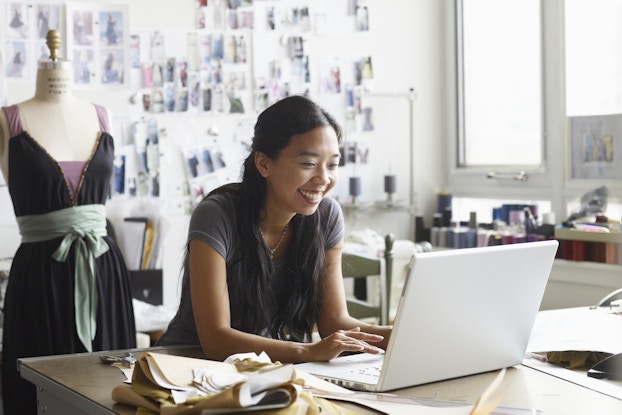  A smiling woman leans on a table in a fashion designer's studio and types on an open laptop. Beside the laptop are folded lengths of fabric in various shades of brown and white. Behind the woman is a wall covered with snapshots of models in dresses and a mannequin dressed in a dark purple dress with a pale mint green sash and lavendar arm straps.