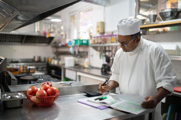  A chef stands in a fully outfitted kitchen and looks down at a handwritten spreadsheet and a calculator. The chef is a man wearing a short white hat and white chef's uniform. He holds a pen in one hand and a piece of paper in the other.