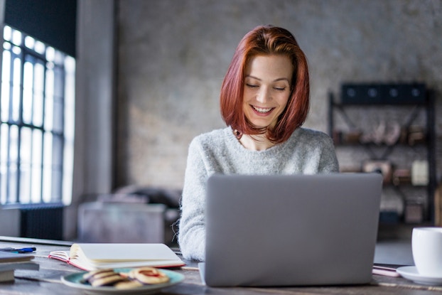 A smiling woman with dyed red hair sits at a table and types on a laptop. Beside the laptop on the table are an open book and a plate of thumbprint cookies. The room behind the woman is a large open space with gray walls, a dark gray sofa, a set of shelves, and a large window letting in bright daylight.