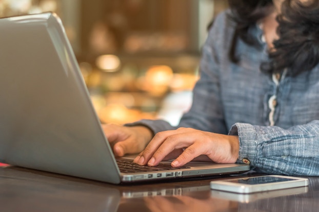 A close-up shot of a person photographed from the shoulders down typing on a laptop. The person has long, dark hair and wears a chambray button-up shirt. The laptop sits open on top of a chrome table. A white smartphone sits screen-up next to it.