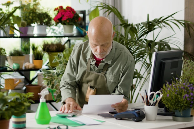  A bald, white-bearded man looks down at the piece of paper in his hand with a serious expression. He leans on a counter in a store filled with potted plants and flowers. Plants of various sizes sit in pots on shelves behind him, on the edges of the counter, and, in the case of a few tall plants, on the floor near the walls. The man wears a muted green collared shirt and a khaki green apron. On the counter in front of him are more folded up pieces of paper, a smart phone, a calculator, a handheld credit card reader, and a neon green spray bottle. A computer monitor and keyboard and a mug filled with pens and scissors are also on the counter.