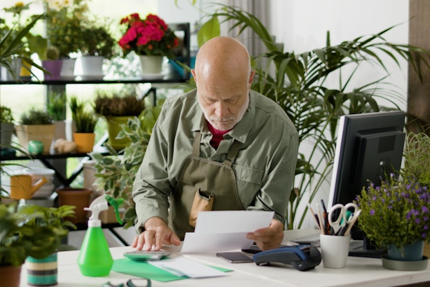 A bald, white-bearded man looks down at the piece of paper in his hand with a serious expression. He leans on a counter in a store filled with potted plants and flowers. Plants of various sizes sit in pots on shelves behind him, on the edges of the counter, and, in the case of a few tall plants, on the floor near the walls. The man wears a muted green collared shirt and a khaki green apron. On the counter in front of him are more folded up pieces of paper, a smart phone, a calculator, a handheld credit card reader, and a neon green spray bottle. A computer monitor and keyboard and a mug filled with pens and scissors are also on the counter.