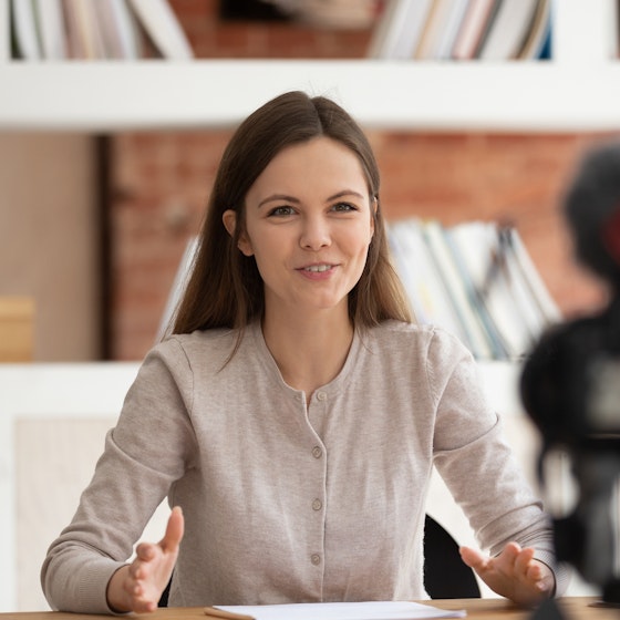 A young woman sits at a desk and speaks to a camera, which sits out of focus in the foreground.