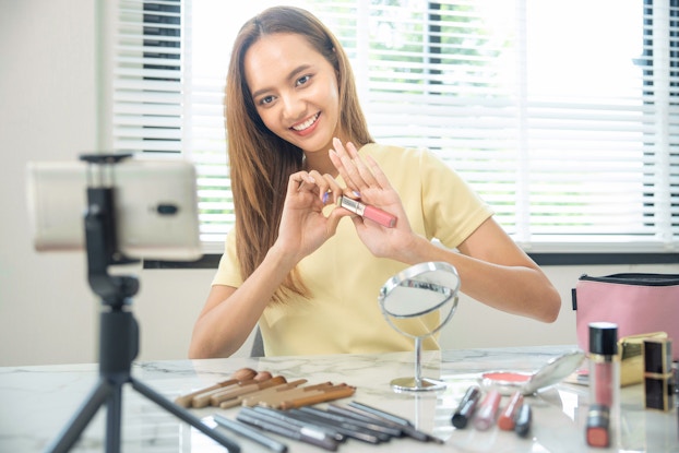 A woman sits at a table, looking and smiling at a smartphone on a tripod in the foreground. The woman holds up a tube of lip gloss for the camera. She has long light brown hair and wears a pale yellow T-shirt. The table in front of her is scattered with more tubes of makeup, makeup brushes, and a small round reversible mirror on a chrome stand.