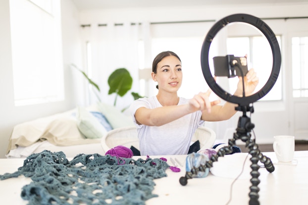  A woman adjusts a smartphone on a camera tripod. On the table next to the camera are piles of yarn and unfinished knitwear.