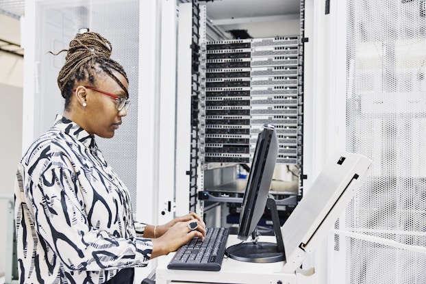 A woman wearing glasses and a white shirt patterned with black wishbone shapes stands in an all-white room and uses a computer, the monitor and keyboard of which are held on an elevated white platform. The woman is seen in profile; past her, on her left, is a doorway, through which can be seen rows of computer servers.