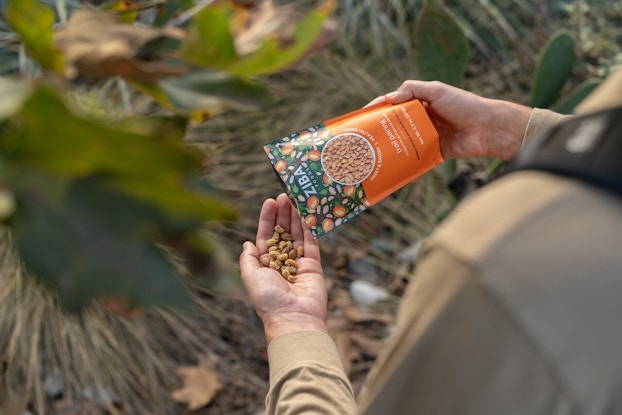 An over-the-shoulder shot of someone pouring Ziba Foods' trail pairing snacks (mulberries and apricot kernals) into their open hand. The Ziba bag is a little over five ounces. Its bottom half is orange and its top half is green and patterned with art of apricot fruits and dried white mulberries.