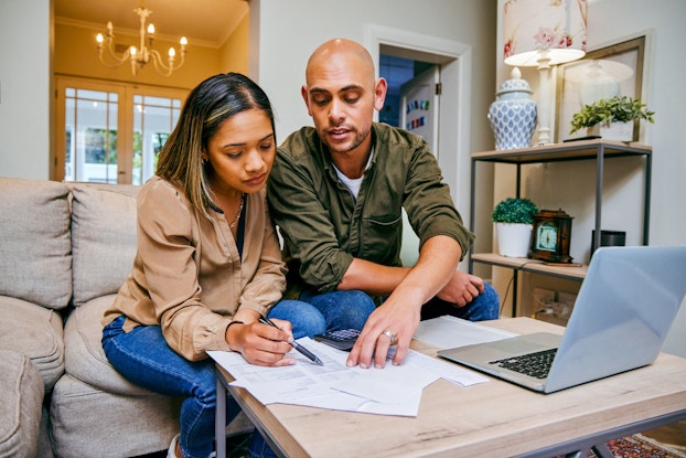 A young couple review their budget in the living room of their home. There are papers spread out on a coffee table next to an open laptop computer.