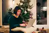 A woman sits on the floor in front of a Christmas tree. On her lap is a laptop on top of a cream-colored pillow. The woman wears a dark green sweater and types on the laptop. On the floor around the woman and the Christmas tree are a few boxes wrapped in silver and gray paper, tied up with string, and decorated with pinecones.